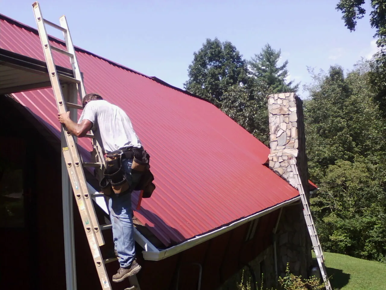 Roofer on a ladder beside a red metal roof and stone chimney — Soffit & Fascia Repair by Roofing Brothers in Opolis